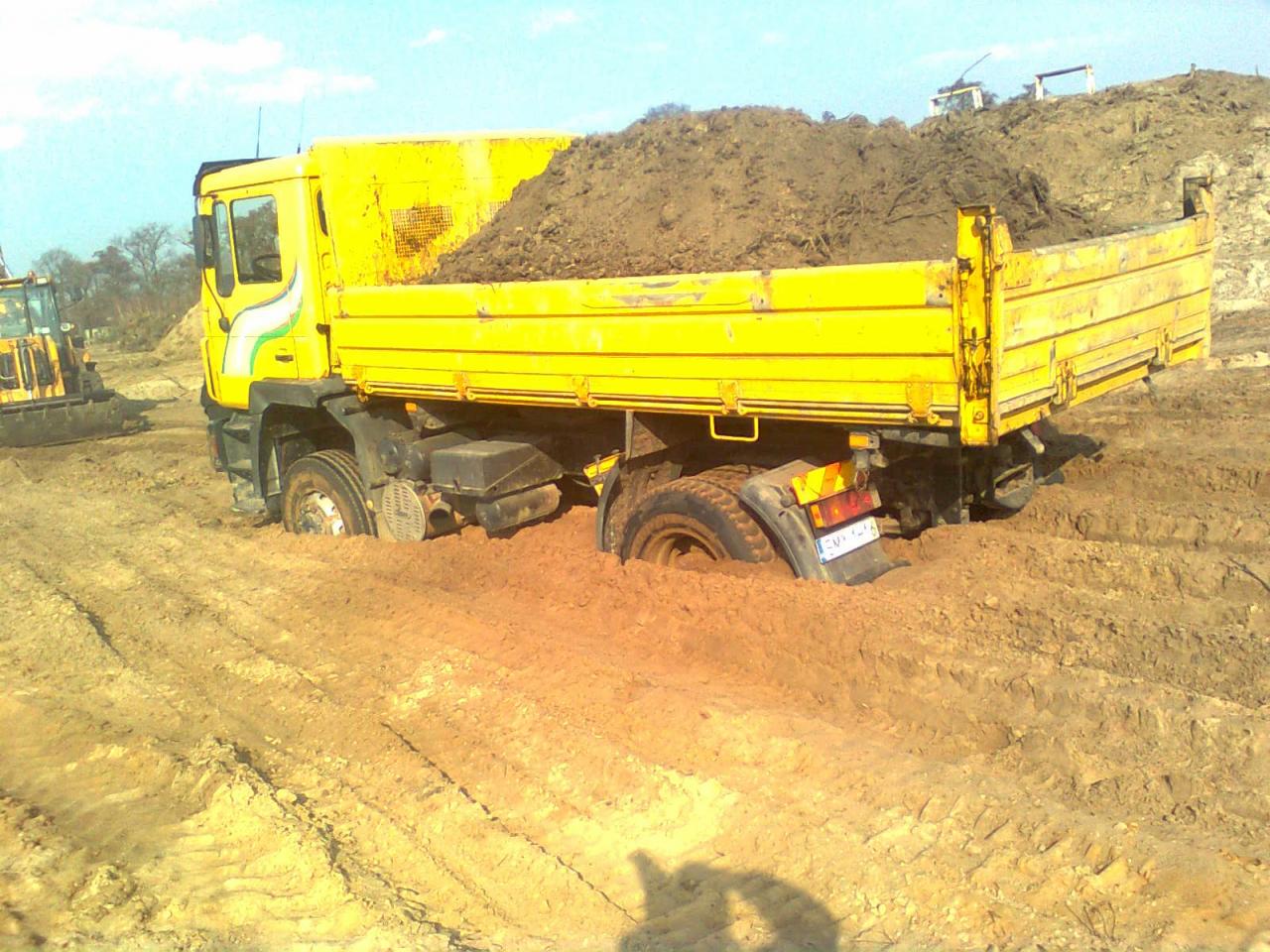 Żółta wywrotka zakopana w błotnistym terenie budowy, załadowana ziemią, z widoczną koparką w tle.