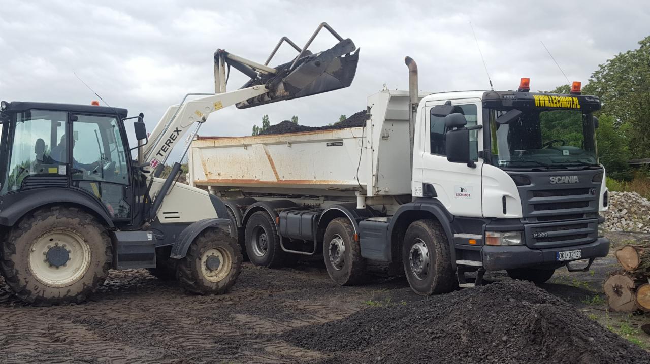 Ładowarka teleskopowa Terex nabiera urobek do wywrotki Scania P360 na placu budowy w pochmurny dzień.