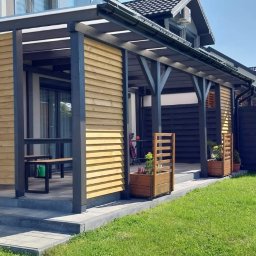 Drew Stol - Zadaszony taras z drewnianymi panelami bocznymi i ciemną konstrukcją nośną, widok z zewnątrz na zadbany trawnik i zabudowę jednorodzinną w tle.