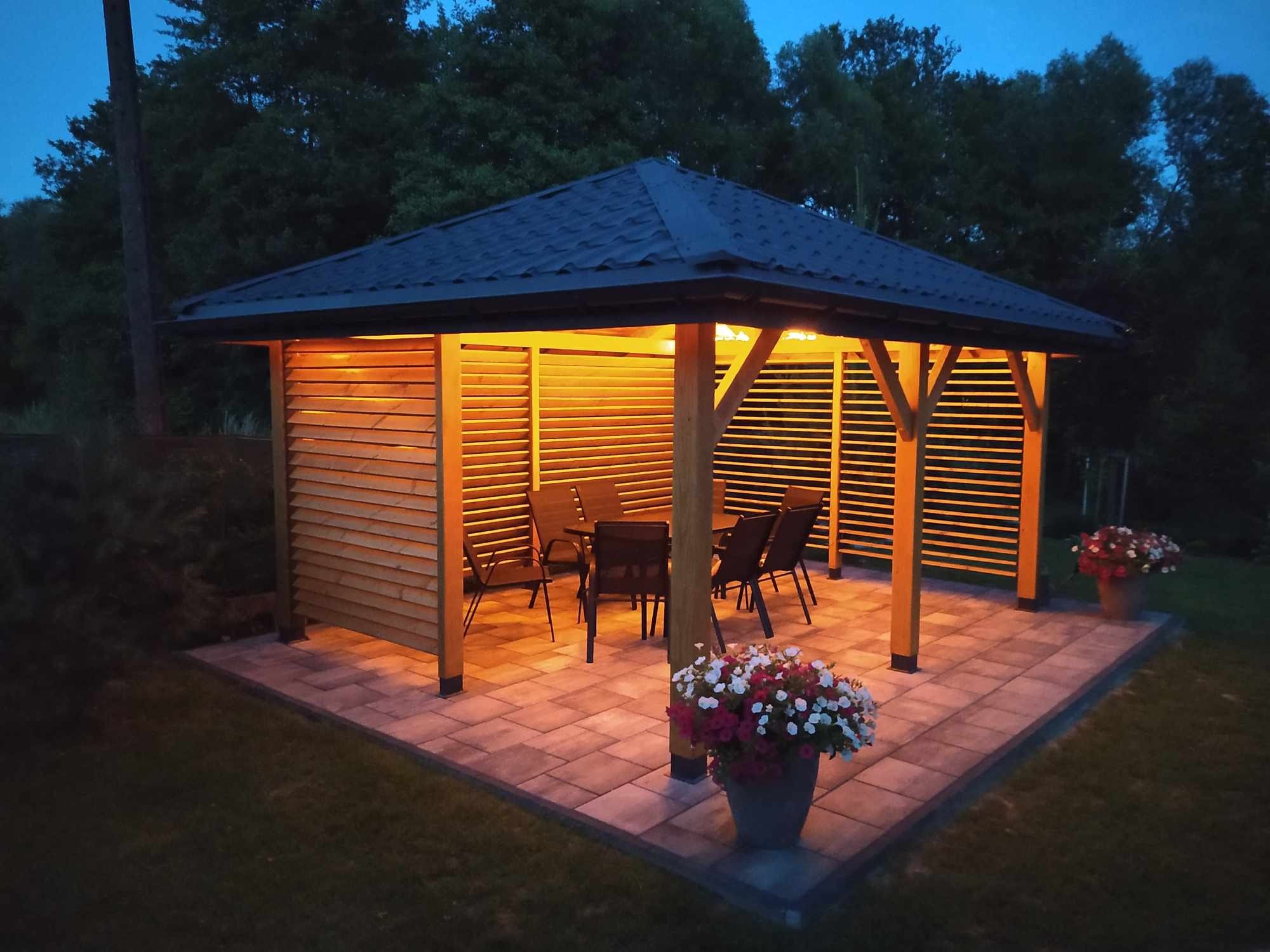 Drewniana altana z oświetleniem wewnętrznym w Błażowej, ustawiona na wybrukowanym tarasie z donicami kwiatów, widoczna z perspektywy ogrodu o zmierzchu.