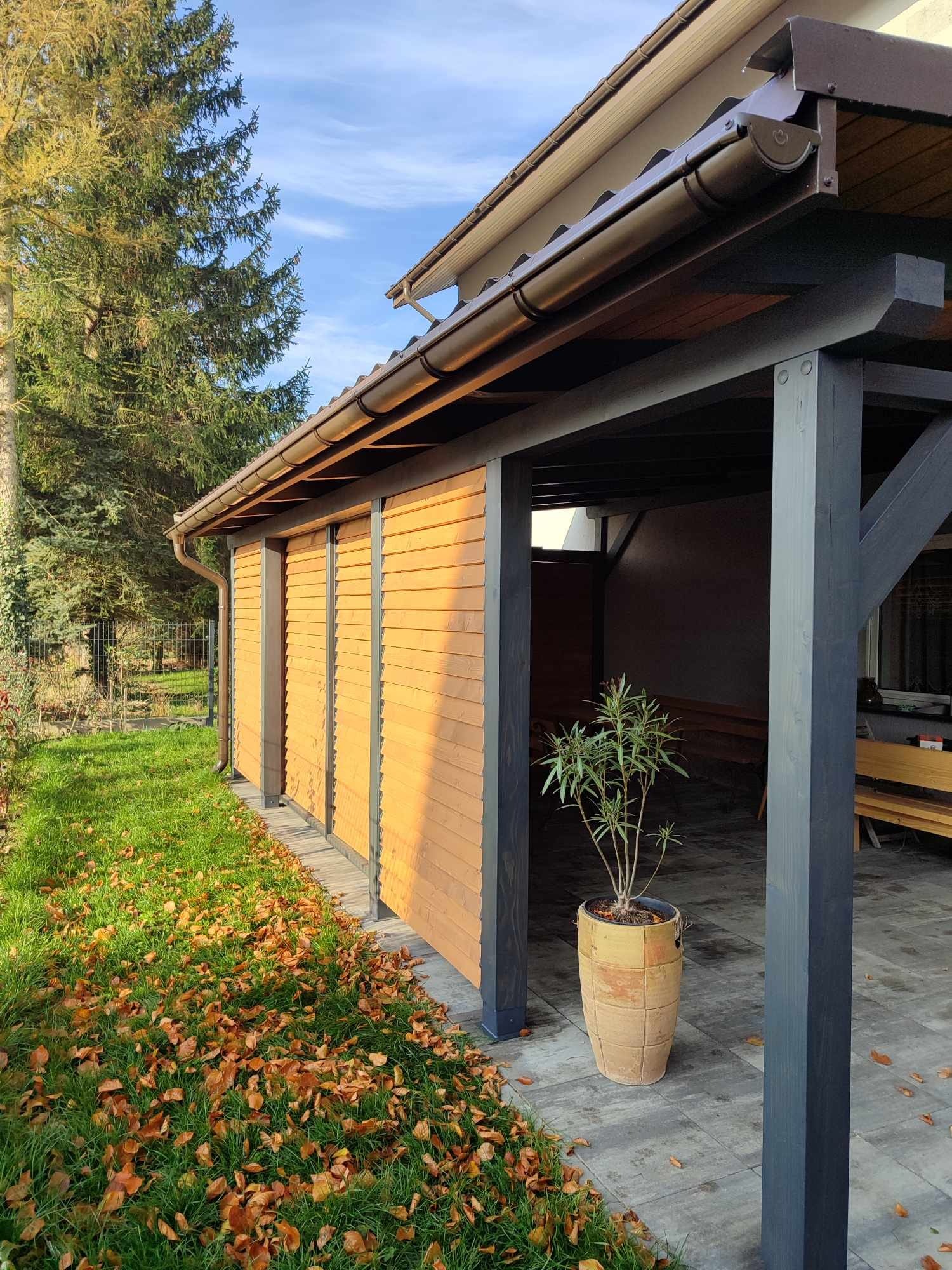 Zadaszona konstrukcja z drewnianymi, żaluzjowymi ścianami bocznymi, ciemnymi słupami nośnymi i rośliną w ozdobnej donicy na szarym bruku, widok z zewnątrz na trawnik pokryty opadłymi liśćmi.