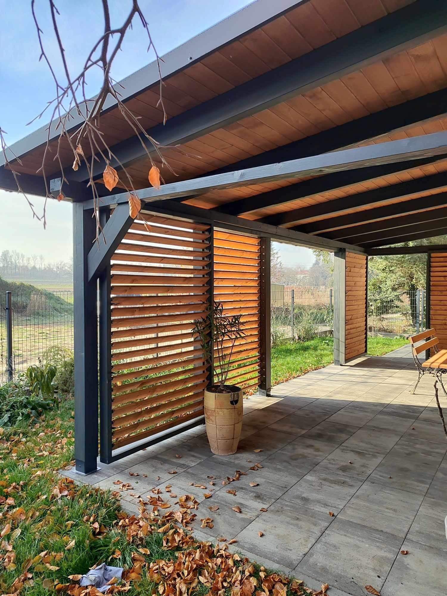 Zadaszony taras z drewnianymi panelami lamelowymi, szarą konstrukcją i kamienną podłogą, widok na ogród przez otwartą przestrzeń, jesienne liście na trawie.