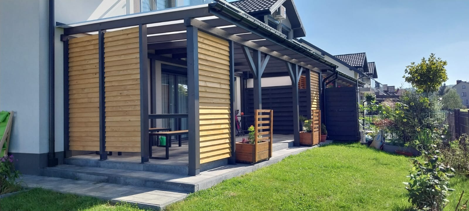 Zadaszony taras z drewnianymi panelami bocznymi i ciemną konstrukcją nośną, widok z zewnątrz na zadbany trawnik i zabudowę jednorodzinną w tle.