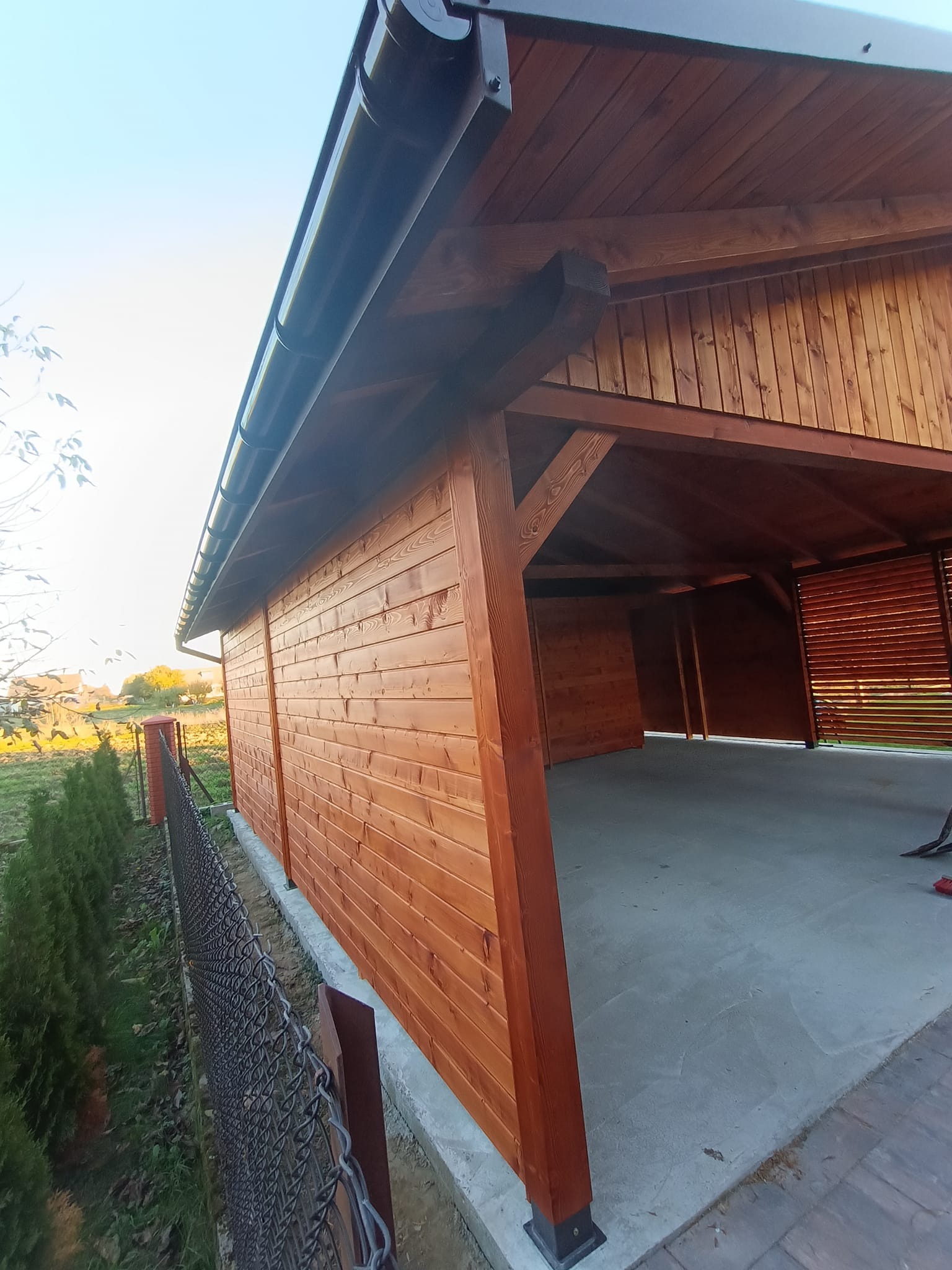 Nowoczesna, drewniana wiata garażowa z brązowymi elementami wykończeniowymi, widok z zewnątrz, rynna odprowadzająca wodę, częściowo ażurowa ściana boczna.