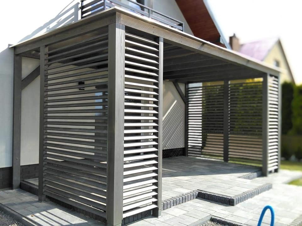 Nowoczesna, zadaszona konstrukcja tarasowa z szarymi, poziomymi listwami, wyłożona szarą kostką brukową, z widokiem na dom z czerwonym dachem w tle.