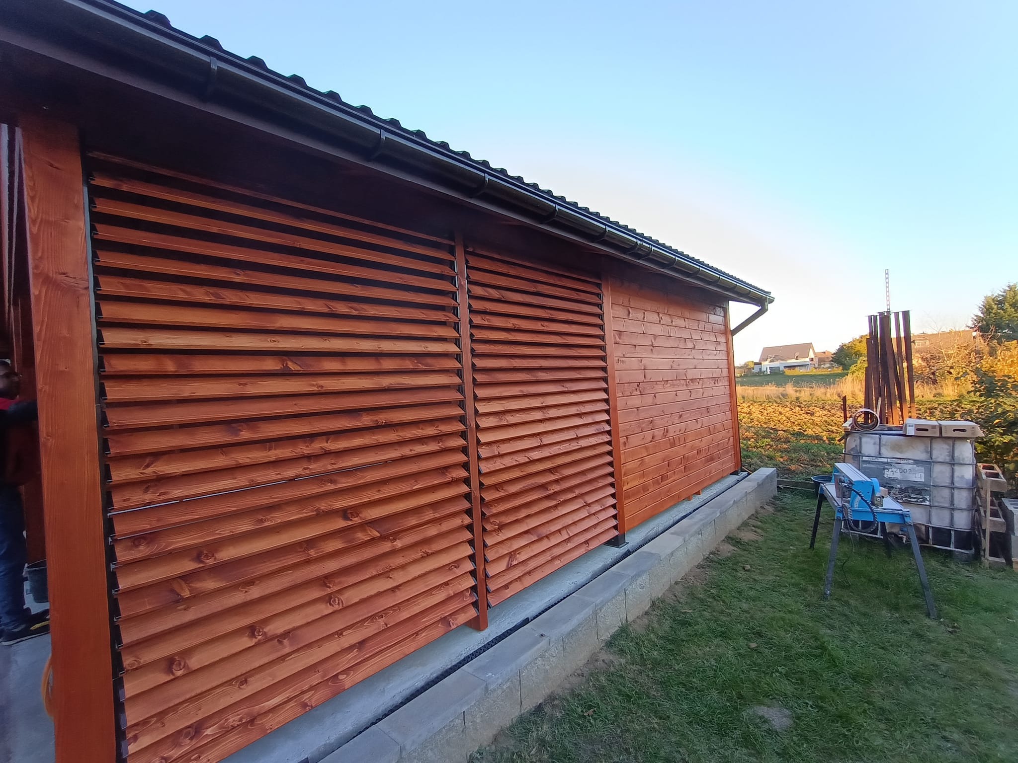 Drewniana konstrukcja z lamelami i deskami w kolorze brązowym, widoczne elementy dachu, narzędzia i materiały budowlane w tle, prawdopodobnie w trakcie montażu lub tuż po nim.