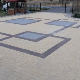 Beżowa kostka brukowa ułożona we wzór z szarymi obramowaniami i kwadratami, widoczny fragment ogrodu z obrzeżem z kostki i drewniane palety w tle.