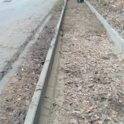 Przygotowanie podłoża pod chodnik z kostki brukowej: żółta koparka i pracownicy wyrównują teren wzdłuż drogi, widoczne krawężniki i opadłe liście.