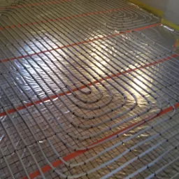 Układanie rur ogrzewania podłogowego na folii izolacyjnej z widocznymi czerwonymi paskami mocującymi