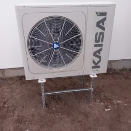 Zewnętrzna jednostka pompy ciepła marki Kaisai, zamontowana na metalowym stelażu na ziemi, przy białej ścianie budynku.