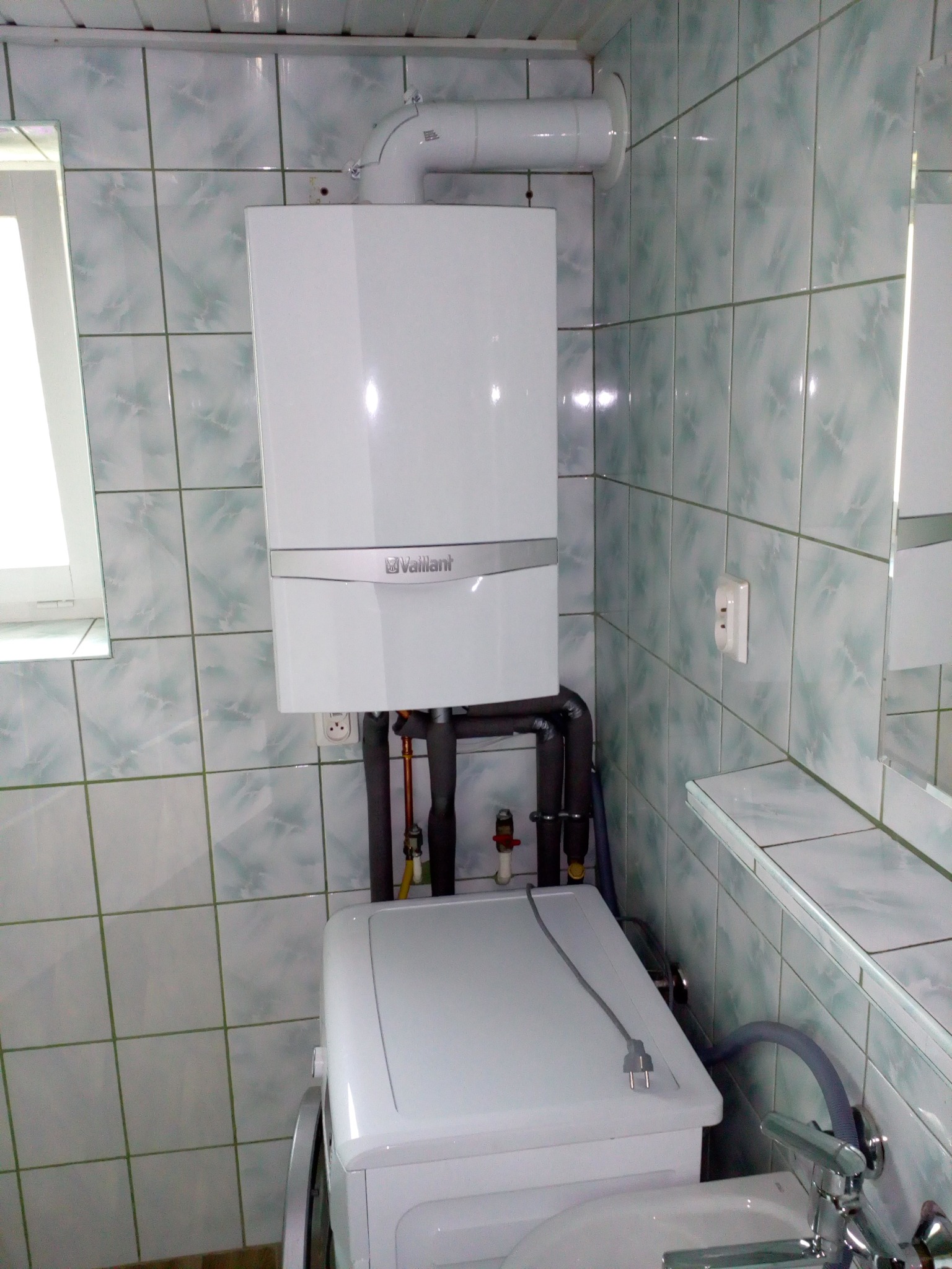 Biały, wiszący piec gazowy marki Vaillant zamontowany w łazience z zielono-białymi płytkami, widoczne rury odprowadzające spaliny i podłączenie do instalacji, poniżej pralka.