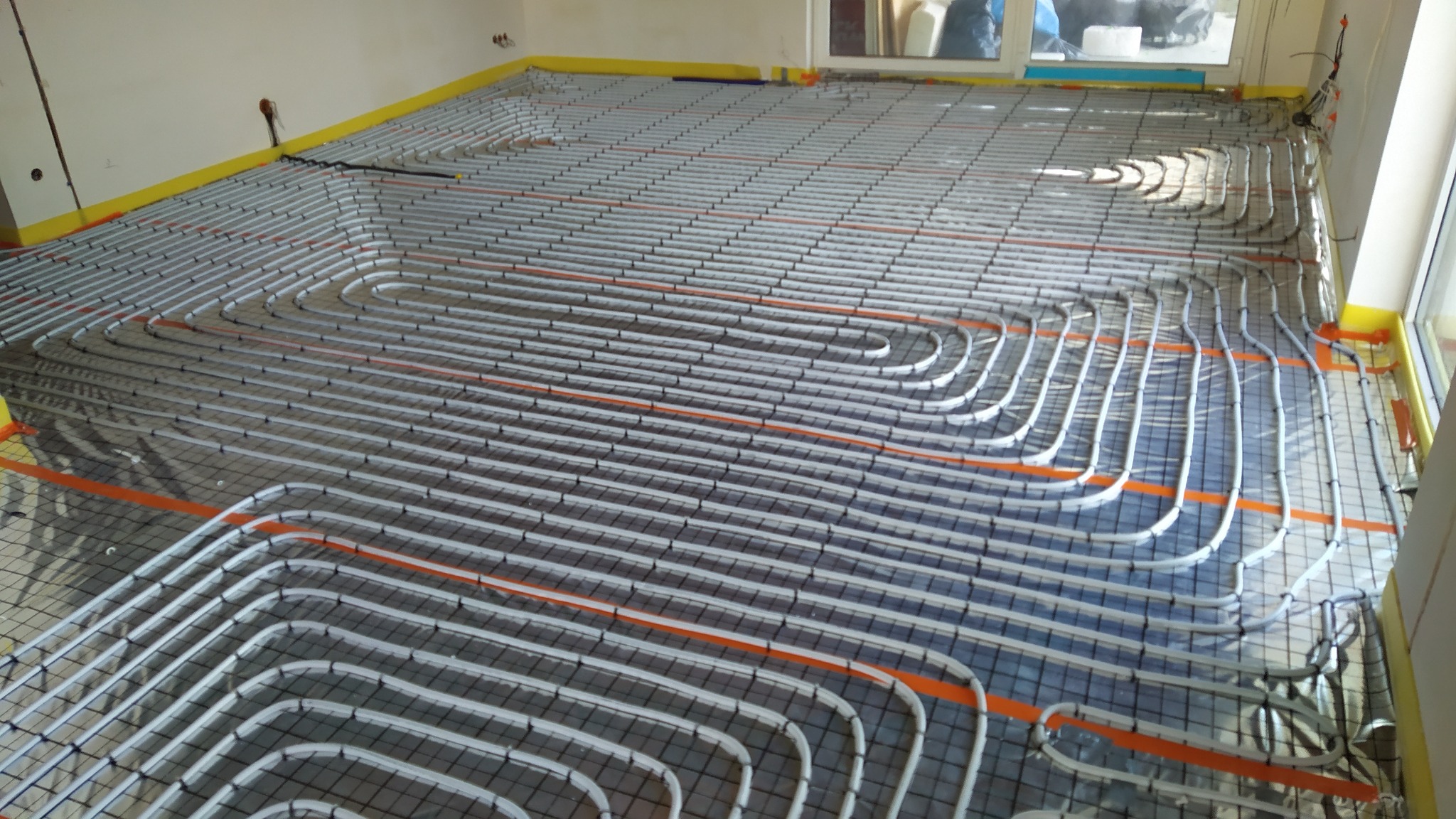 Rozłożone rury ogrzewania podłogowego na metalowej siatce, widoczne pomarańczowe taśmy mocujące, żółta folia przy ścianach, surowy stan wykończenia wnętrza.