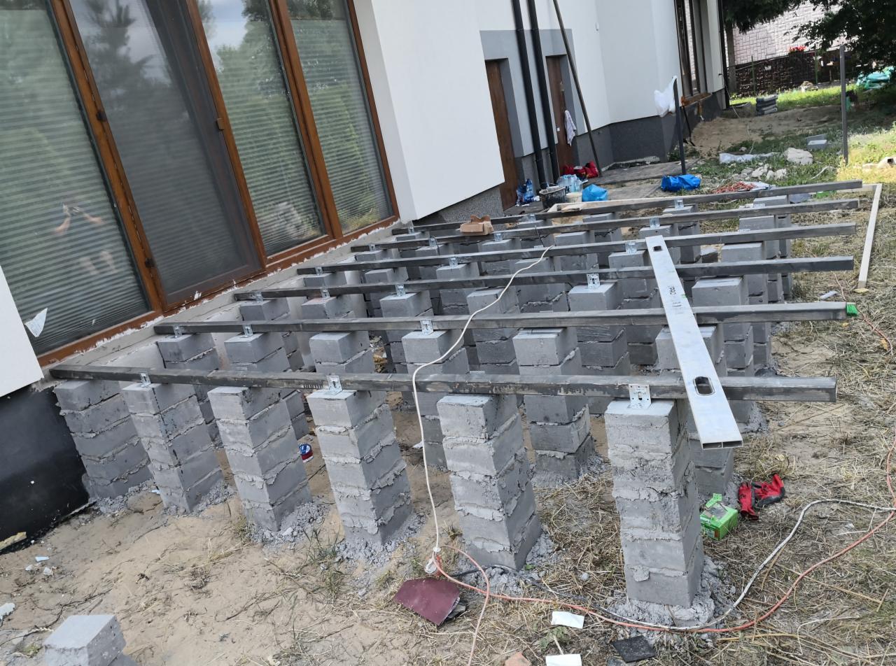 Konstrukcja nośna tarasu z bloczków betonowych i metalowych belek, widoczne poziomica, przewody i narzędzia, etap budowy.