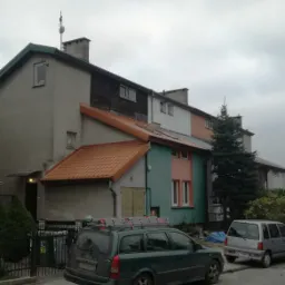 Widok na szereg domów jednorodzinnych o zróżnicowanej architekturze i kolorystyce elewacji, z pomarańczowym dachem na jednym z budynków. Na dachu zaparkowanego samochodu drabina dekarska.