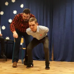 Dwóch mężczyzn na drewnianej scenie przed niebieską zasłoną w białe gwiazdki, jeden w koszuli w kratę i dżinsach, drugi w jasnej koszuli z czerwonym sercem i ciemnych spodniach, w trakcie występu...