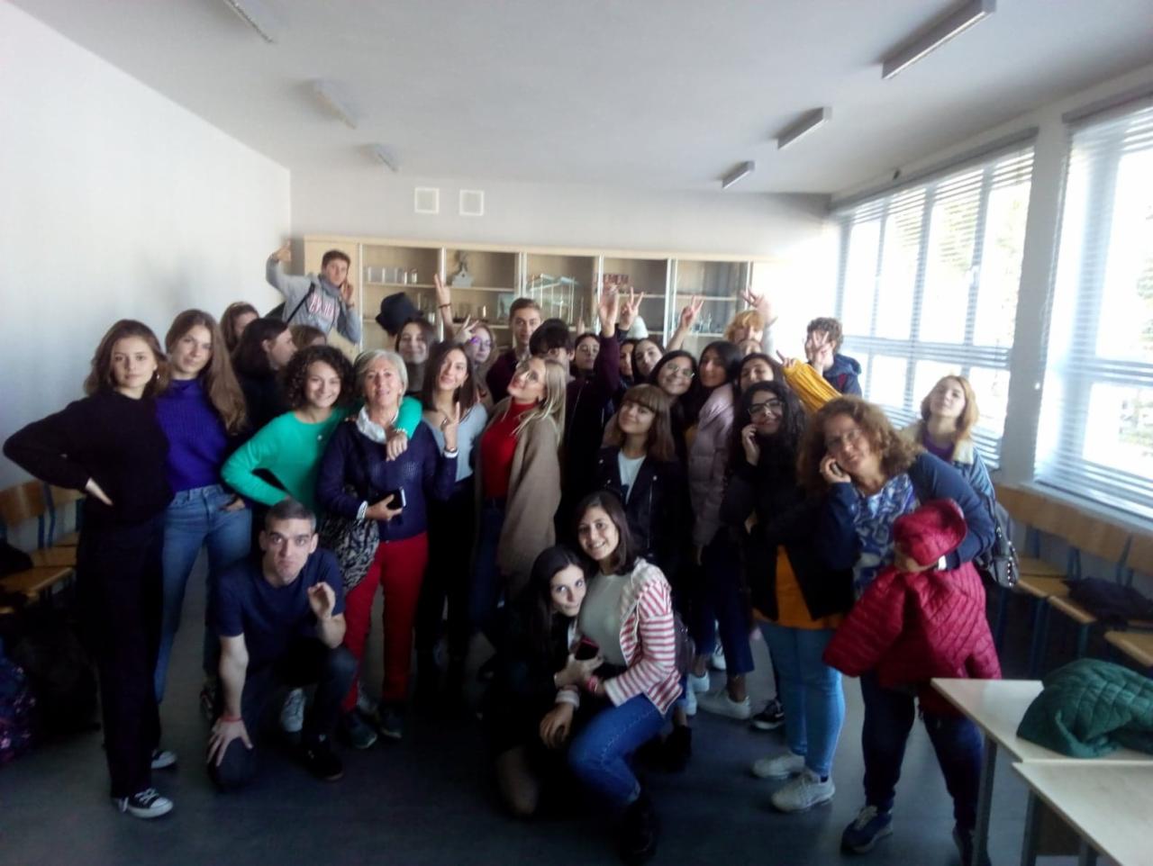 Duża grupa uczniów i nauczycieli pozuje do zdjęcia w klasie z białymi ścianami i oknami z żaluzjami.