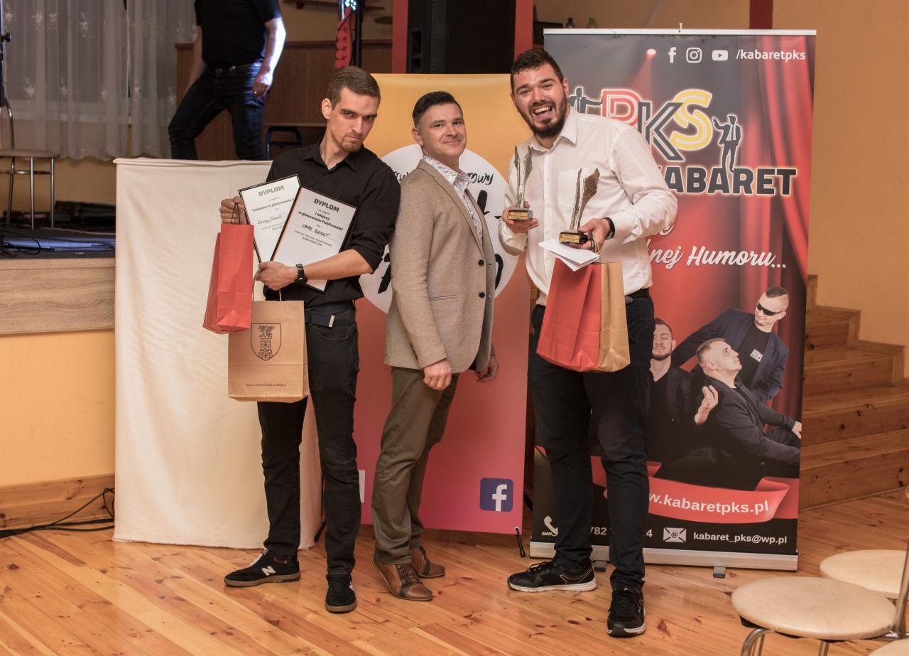 Trzech mężczyzn pozuje z dyplomami i statuetkami na tle baneru reklamowego kabaretu PKS.