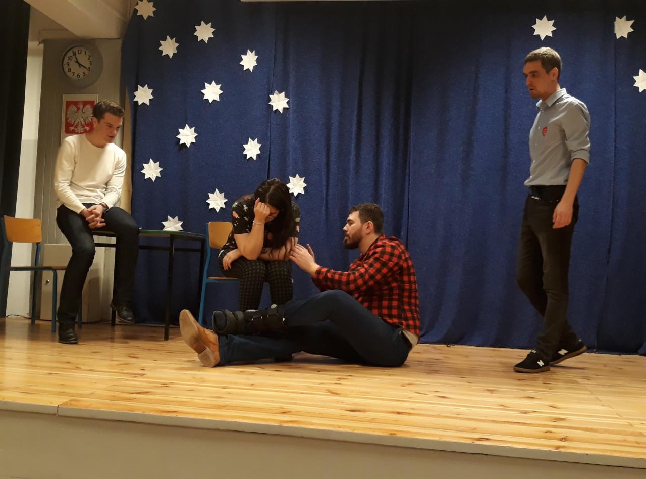 Scena z przedstawienia: aktor w czerwonej koszuli w kratę pociesza aktorkę siedzącą na krześle, w tle dekoracja z granatowej tkaniny z białymi gwiazdami, dwóch aktorów w tle.