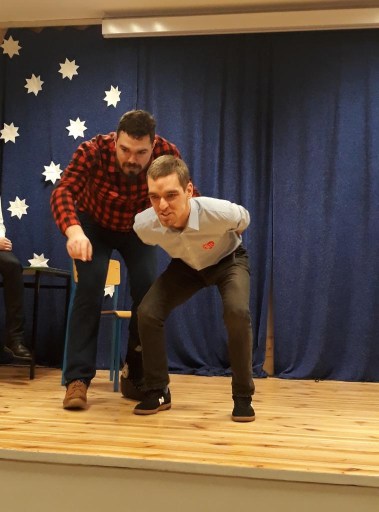 Dwóch mężczyzn na drewnianej scenie przed niebieską zasłoną w białe gwiazdki, jeden w koszuli w kratę i dżinsach, drugi w jasnej koszuli z czerwonym sercem i ciemnych spodniach, w trakcie występu...