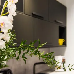 Nowoczesna kuchnia z czarnymi szafkami, białym blatem i dekoracyjnym bukietem białych orchidei na pierwszym planie, żółty kubek widoczny w otwartej półce.