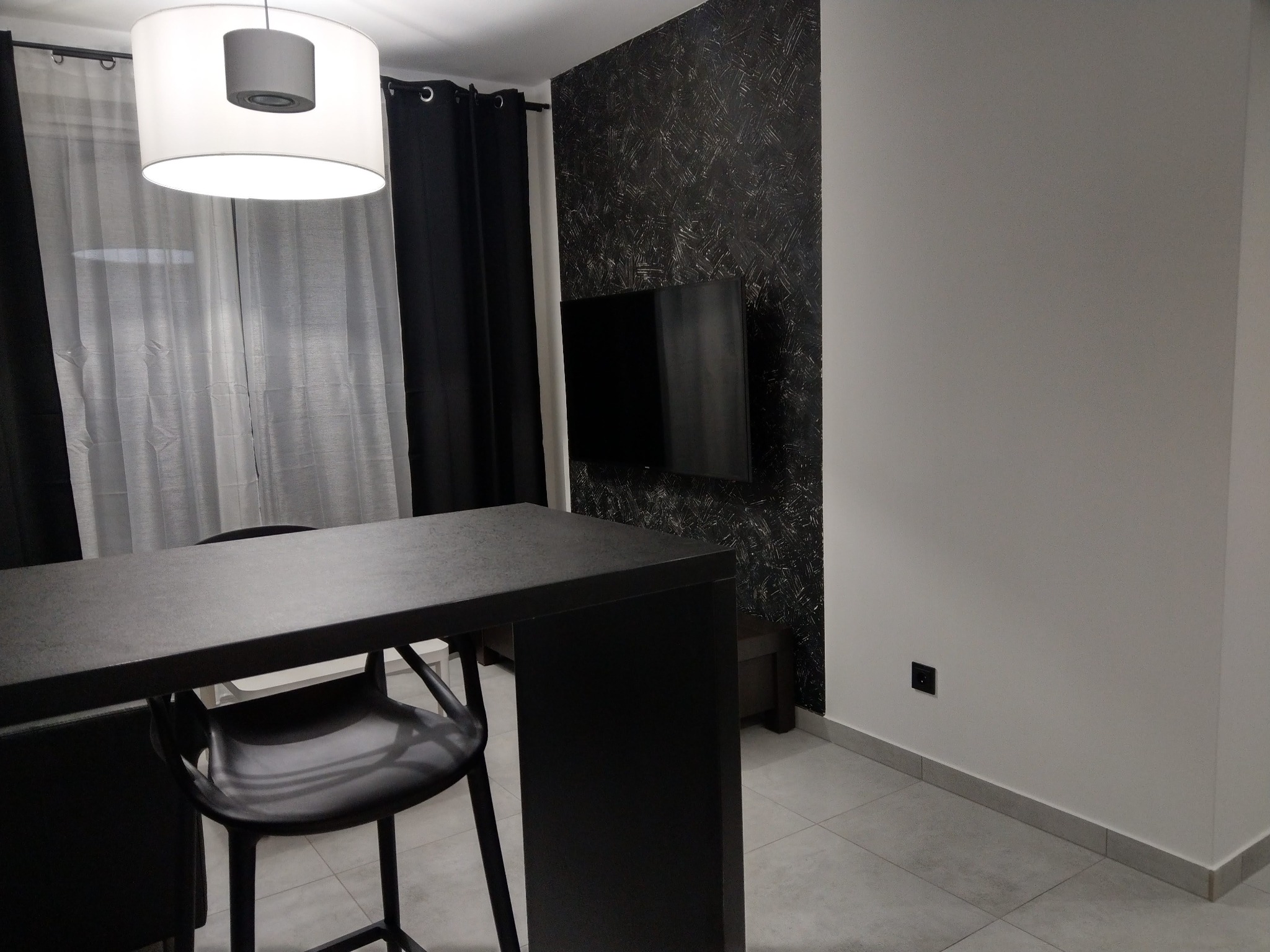 Wnętrze pokoju z czarnym stołem barowym, krzesłem, telewizorem na ścianie z dekoracyjną fakturą, białą ścianą i szarymi płytkami podłogowymi, oświetlone okrągłą lampą wiszącą.