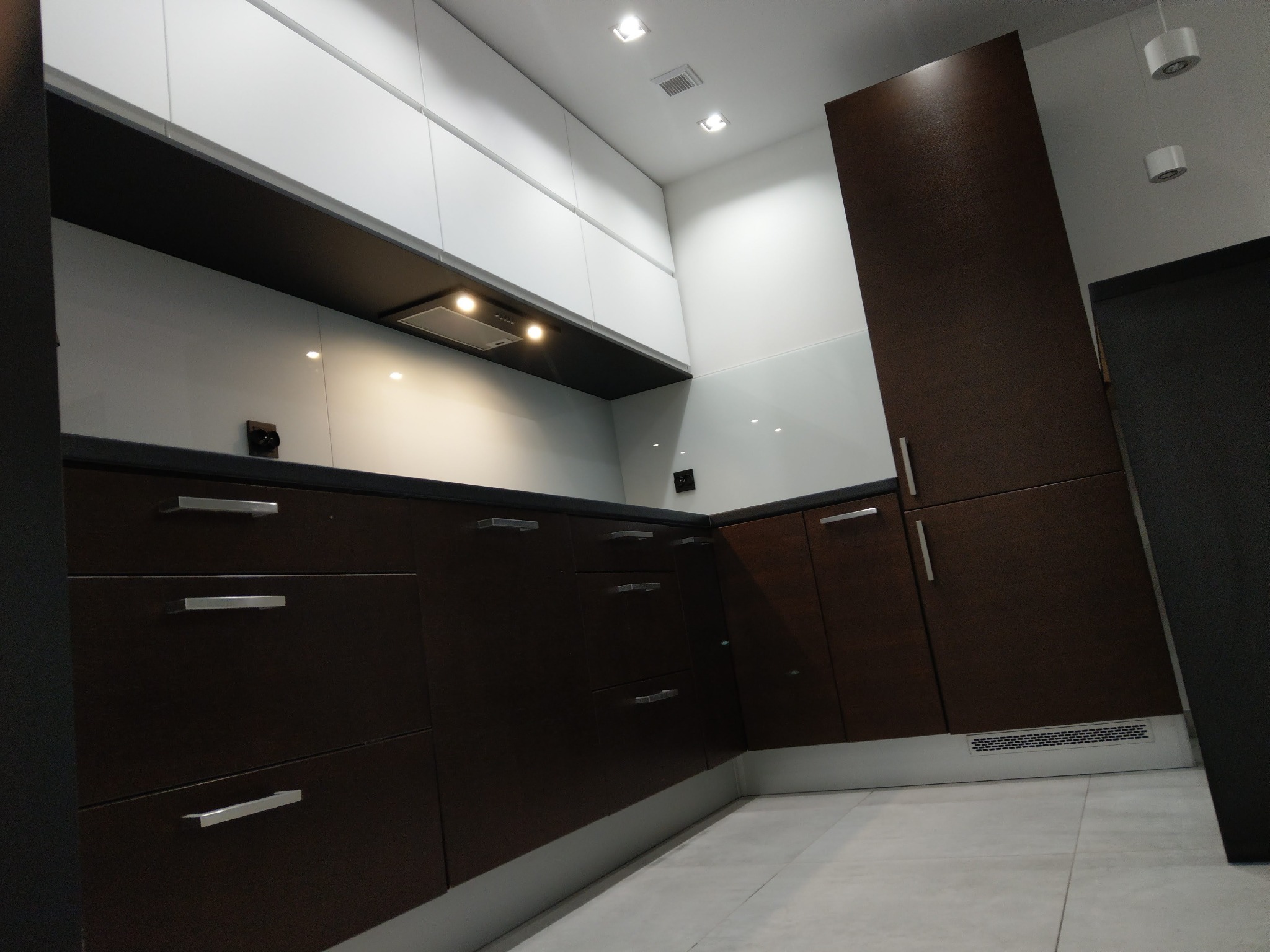 Nowoczesna kuchnia w brązach i bieli z połyskującymi frontami, oświetleniem punktowym i szarymi płytkami podłogowymi, uchwycona pod kątem z niskiej perspektywy.