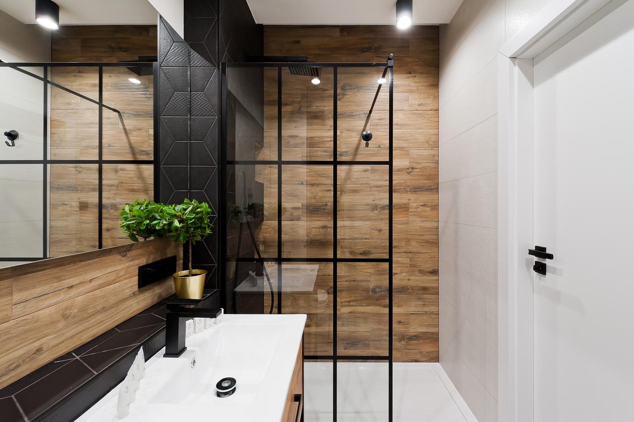 Nowoczesna łazienka z kabiną prysznicową typu walk-in z czarnymi profilami, drewnopodobnymi płytkami na ścianach i dekoracyjną ścianą z czarnych, geometrycznych płytek, umywalką z czarną armaturą...
