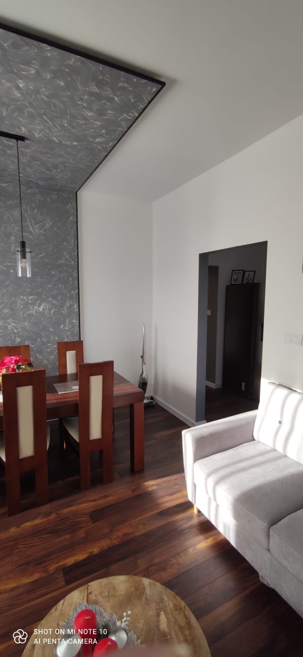 Jasne wnętrze pokoju z drewnianą podłogą, stołem i krzesłami, szarym sufitem z czarną ramą, białymi ścianami i szarą sofą; widoczny odkurzacz pionowy.