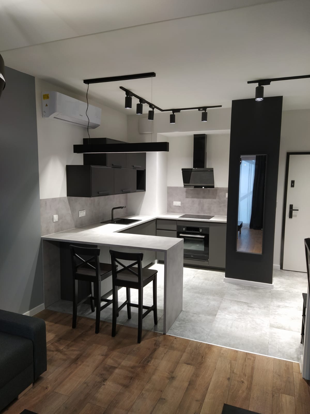 Nowoczesna, szaro-czarna kuchnia z wyspą i hokerami w Jadownikach, oświetlona reflektorami sufitowymi na szynoprzewodach.