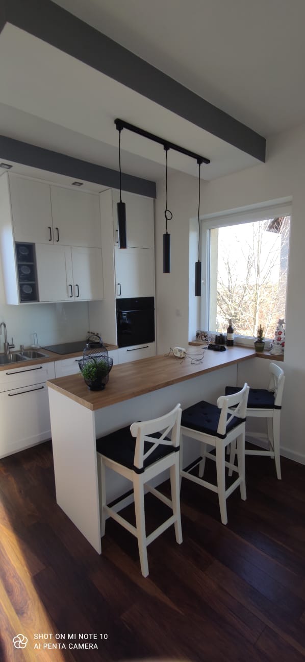 Jasna kuchnia z drewnianym blatem, wyspą kuchenną z trzema białymi hokerami z czarnymi poduszkami, minimalistyczne czarne lampy wiszące nad wyspą, widok na okno z dekoracjami.