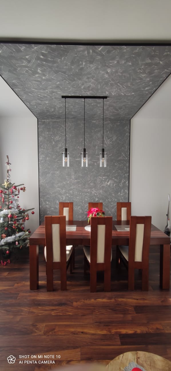 Jadalnia z drewnianym stołem i krzesłami, sufit z dekoracyjną szarą fakturą, wisząca lampa z trzema szklanymi kloszami, parkiet w odcieniach brązu, w rogu choinka.