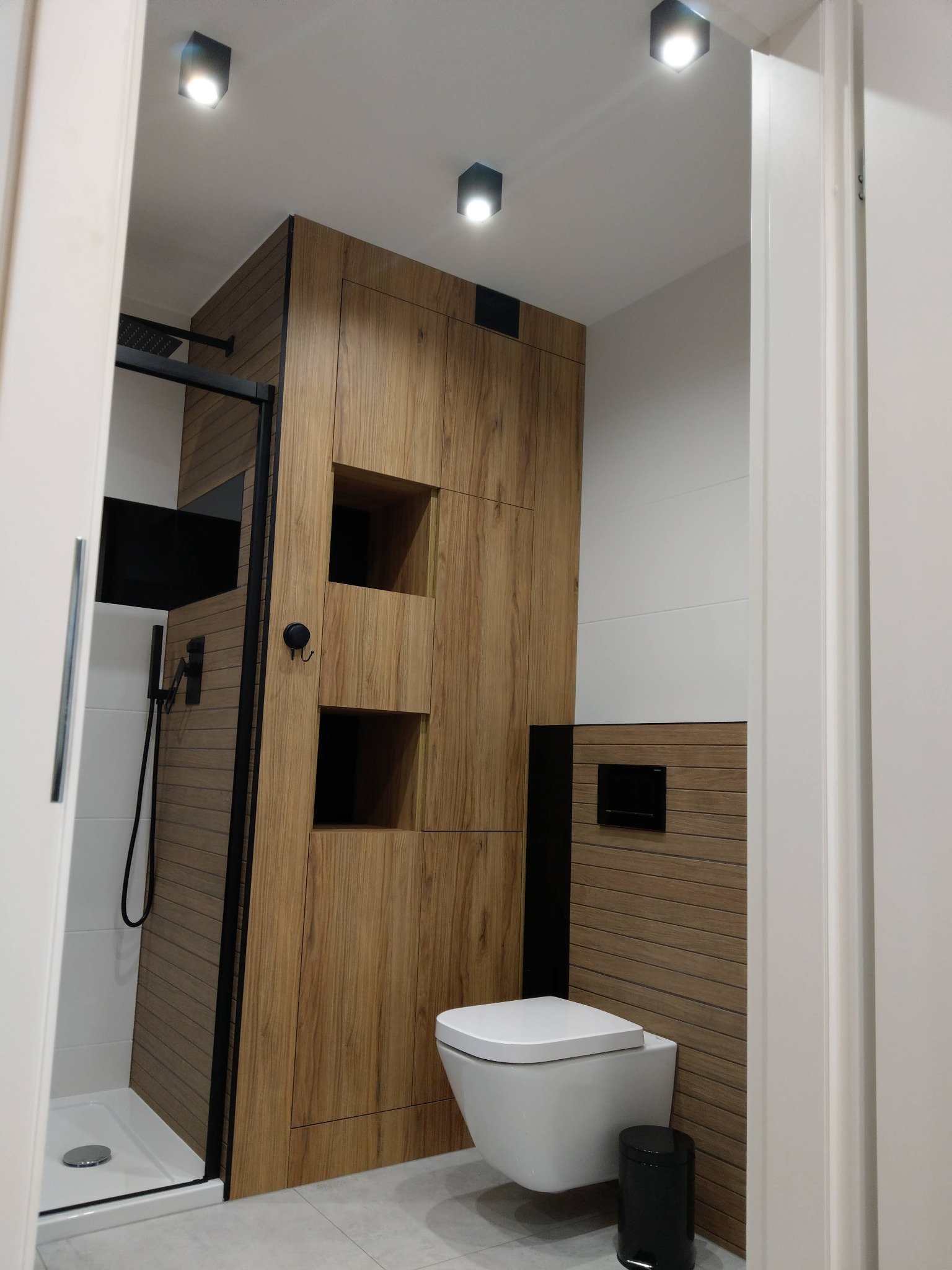 Nowoczesna łazienka z kabiną prysznicową typu walk-in, zabudową z drewnopodobnych paneli z otwartymi półkami, podwieszaną toaletą i czarnymi akcentami w postaci armatury i kosza na śmieci.