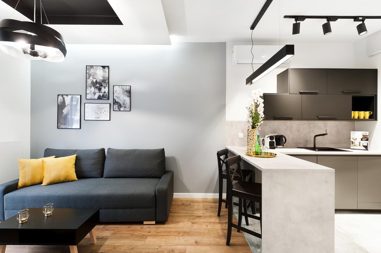 Nowoczesne wnętrze mieszkania z szarą sofą z żółtymi poduszkami, czarnym stolikiem kawowym, czterema obrazami na ścianie, drewnianą podłogą i aneksem kuchennym z barkiem oraz czarnym oświetleniem...