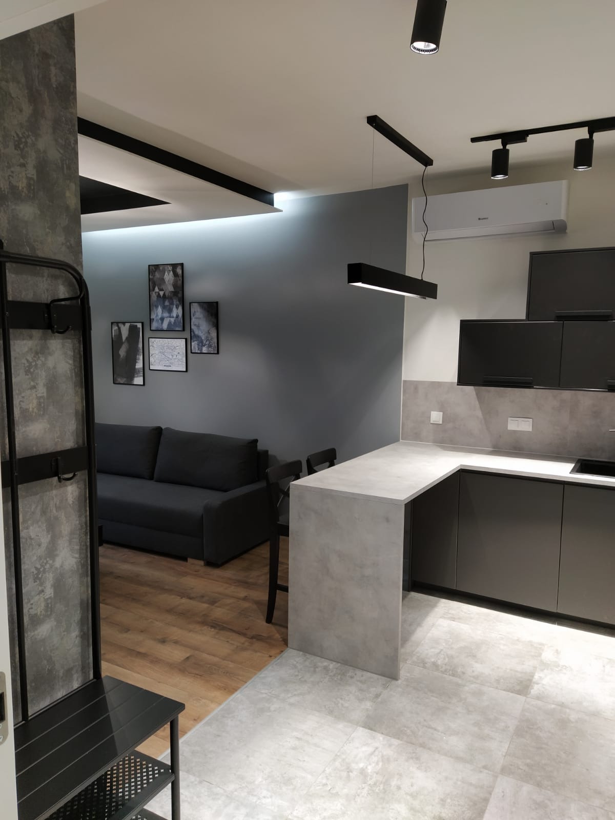Nowoczesne wnętrze mieszkania z szarą sofą, grafitową zabudową kuchenną z blatem imitującym beton, drewnianą podłogą i szarą podłogą z płytek, oświetlone czarnymi lampami sufitowymi.