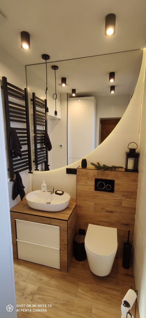 Nowoczesna łazienka z owalną umywalką nablatową, drewnianą zabudową, podświetlanym lustrem o nieregularnym kształcie i czarnym grzejnikiem drabinkowym.