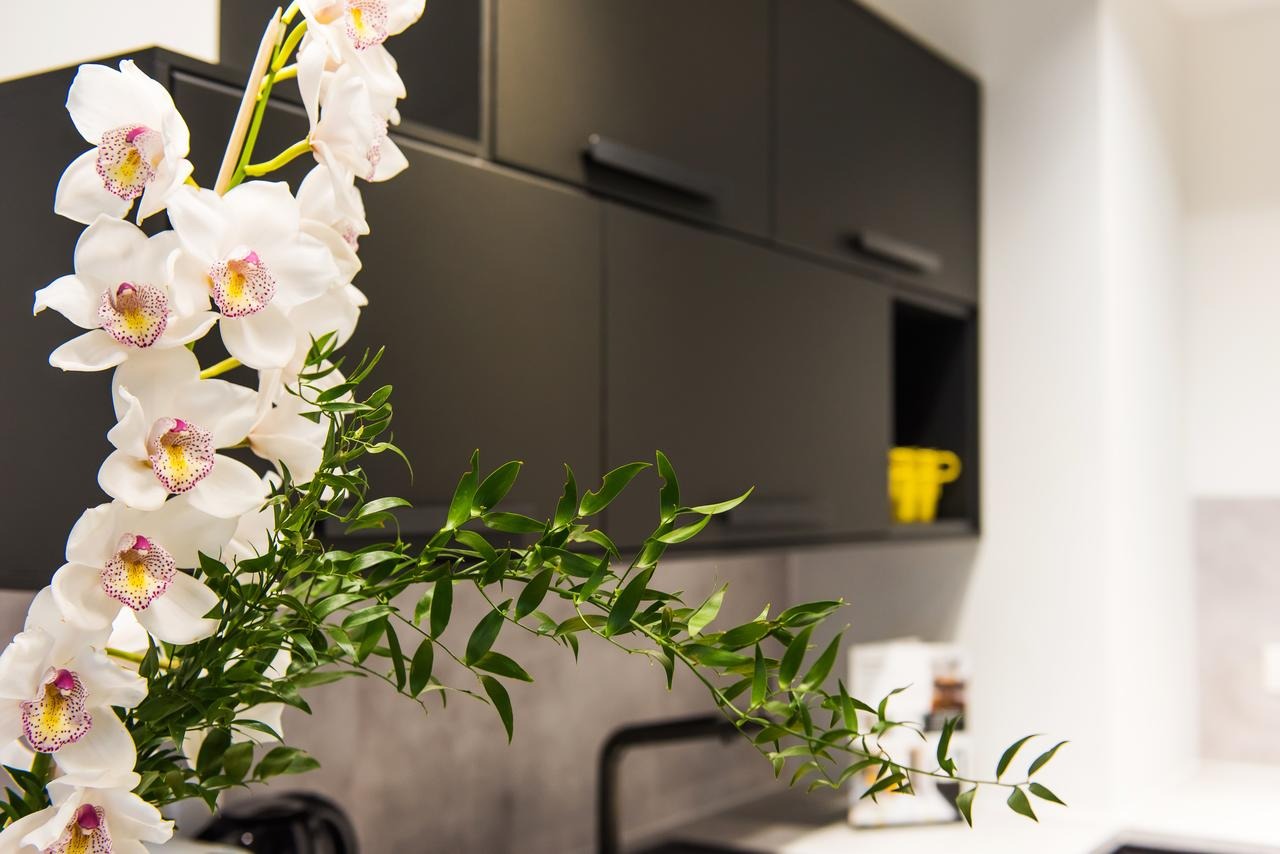 Nowoczesna kuchnia z czarnymi szafkami, białym blatem i dekoracyjnym bukietem białych orchidei na pierwszym planie, żółty kubek widoczny w otwartej półce.
