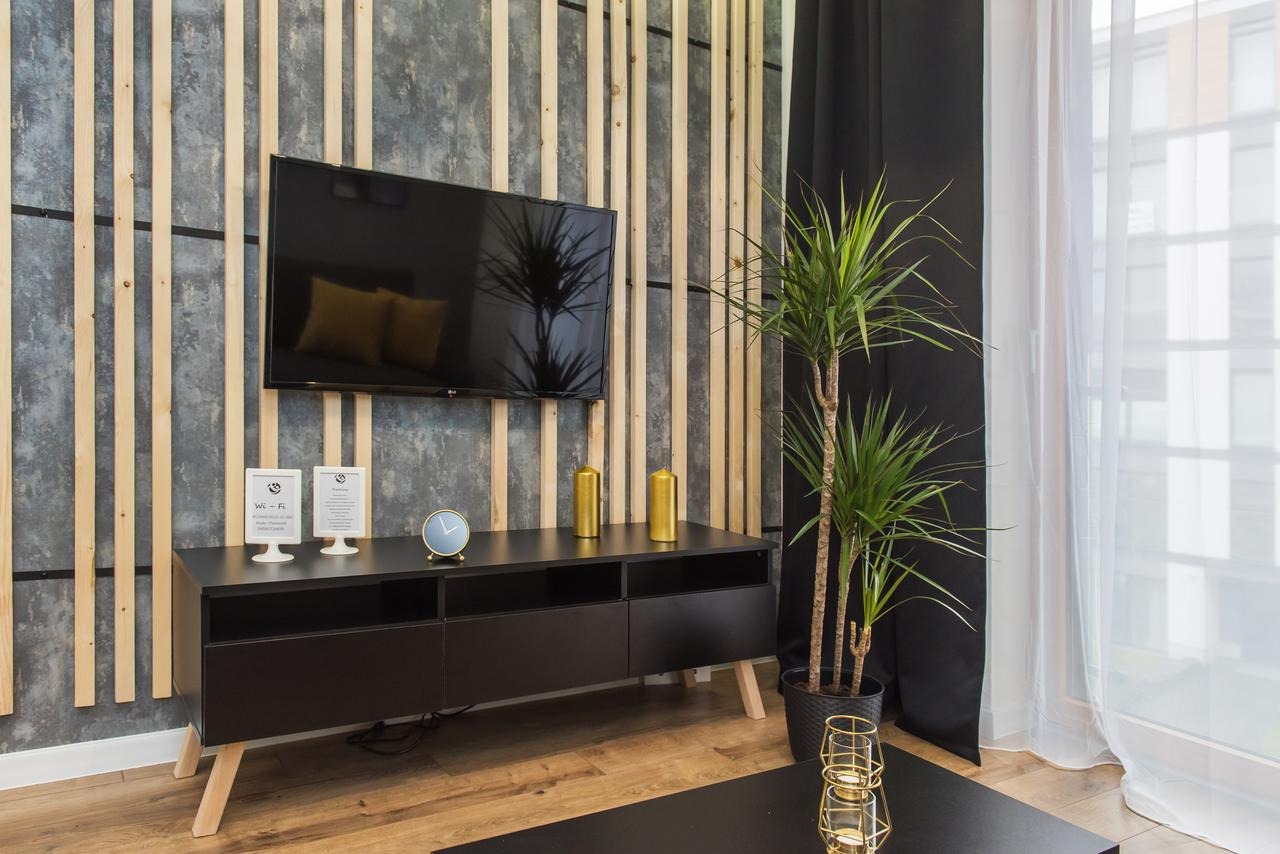 Nowoczesny salon z drewnianą ścianą, telewizorem, czarną szafką RTV na drewnianych nóżkach, zegarem, złotymi świecznikami, rośliną doniczkową i zasłonami.