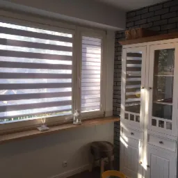 Pokój z oknami osłoniętymi poziomymi przesłonami okiennymi, na parapecie dekoracje, obok biały kredens na tle ściany z cegły.