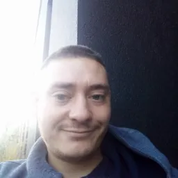 Portret uśmiechniętego mężczyzny z krótkimi włosami na tle ściany w dwóch kolorach. Ubrany w bluzę z kapturem, z widocznym fragmentem okna po lewej stronie.