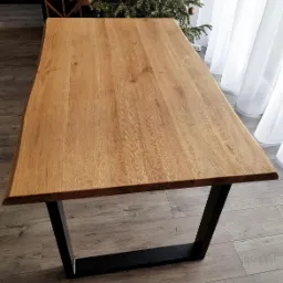 Stół dębowy z blatem o naturalnym wykończeniu, osadzony na czarnych, metalowych nogach w kształcie trapezu, na tle parkietu i zasłon.