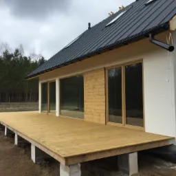 Nowoczesny dom z rozległym tarasem z desek modrzewiowych, widoczne betonowe podpory, okna tarasowe i elewacja częściowo wykończona drewnem.