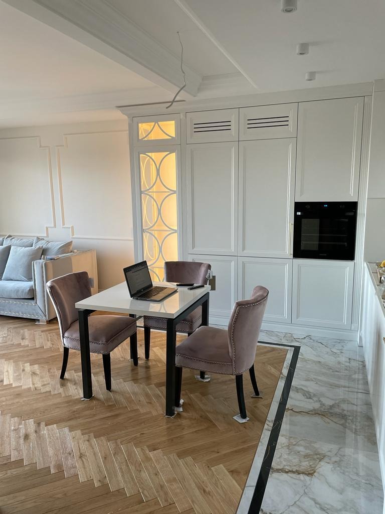 Wnętrze z parkietem ułożonym we wzór jodełki klasycznej, jasne drewno kontrastuje z marmurową posadzką, w tle biała zabudowa kuchenna i stół z krzesłami.