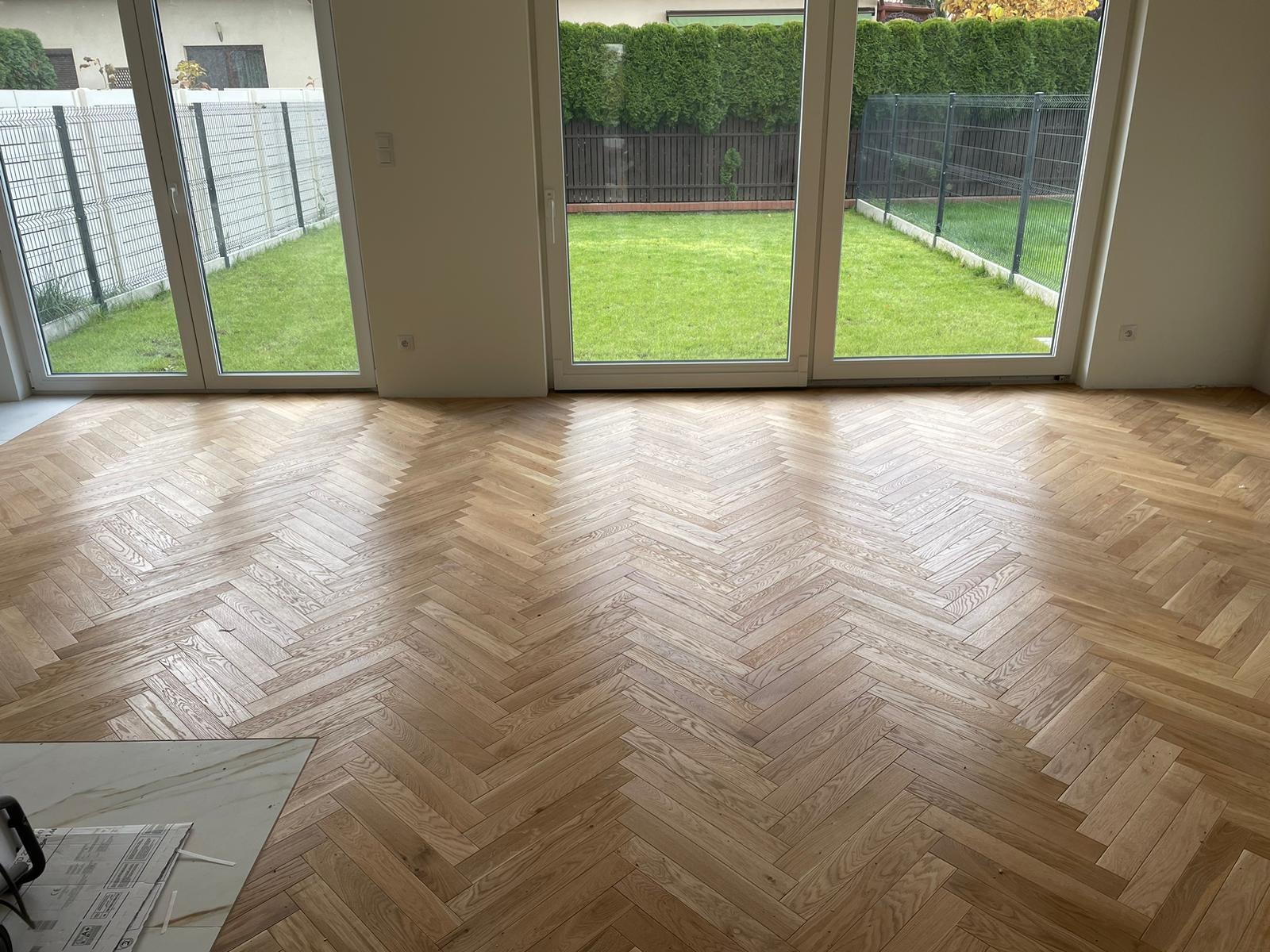 Jasna, dębowa podłoga ułożona w klasyczną jodełkę, widok na ogród przez duże okna tarasowe, częściowo widoczne dokumenty na podłodze w lewym dolnym rogu.