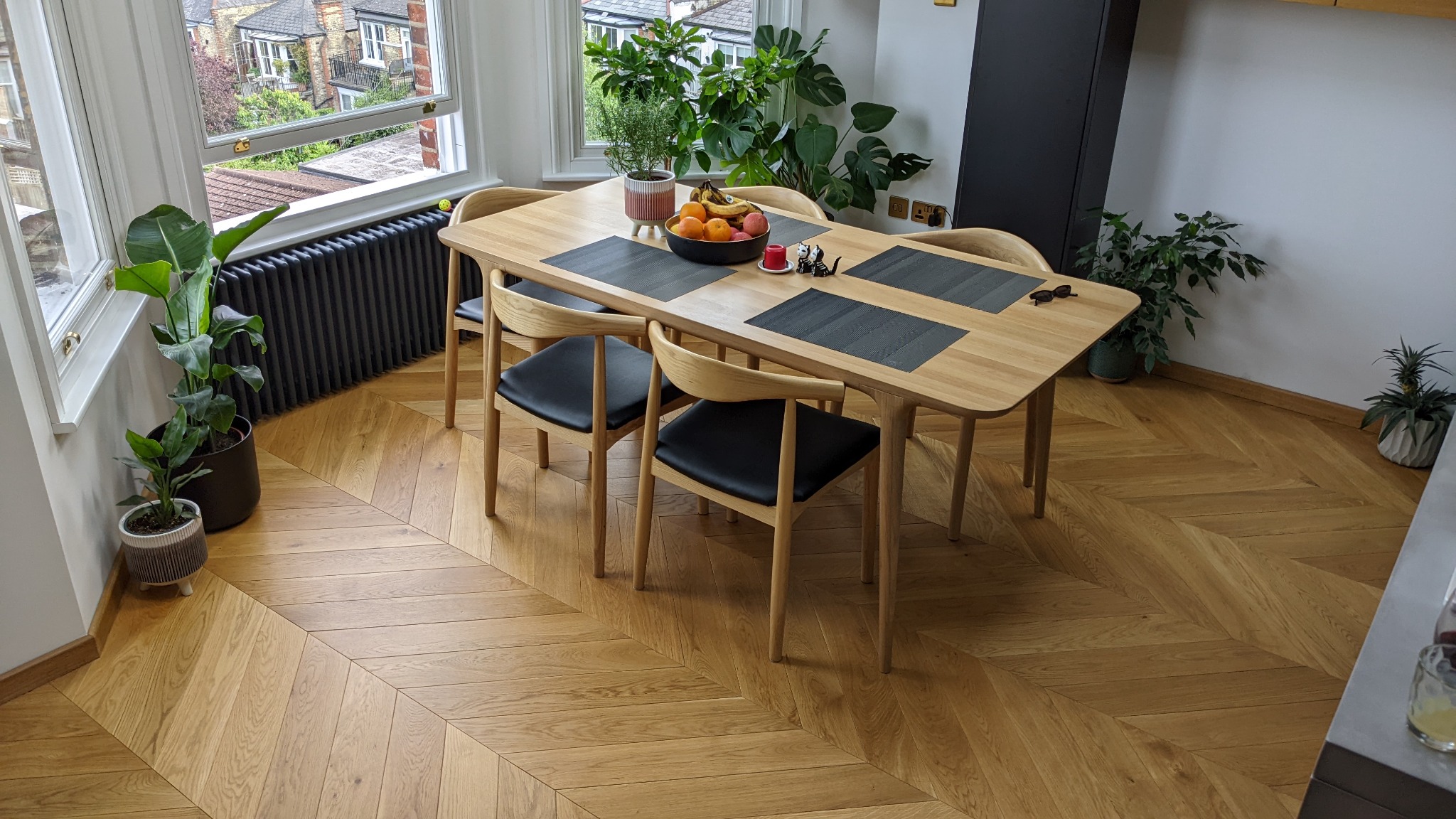 Jasna podłoga dębowa ułożona we wzór jodełki francuskiej w jadalni, widoczne czarne krzesła z jasnego drewna i stół z owocami, rośliny doniczkowe w tle.