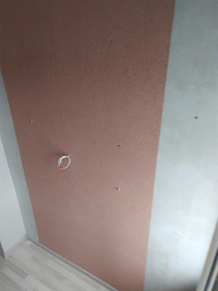 Fragment ściany z nierównomiernie nałożoną, brązową strukturą, widoczne wystające przewody elektryczne i niedokładne wykończenie przy podłodze.