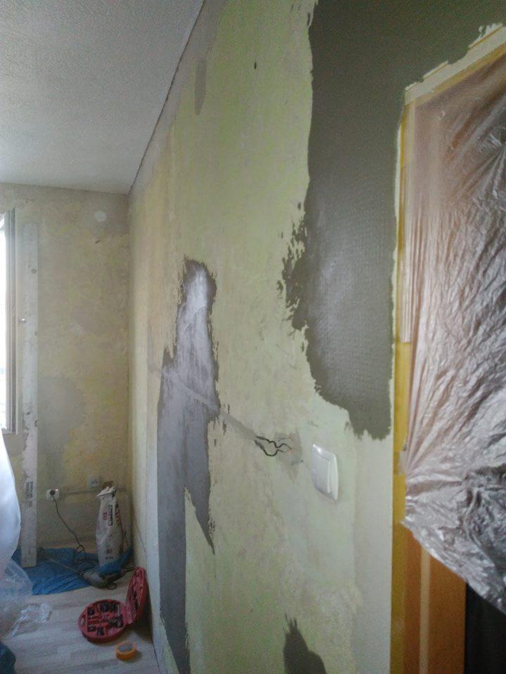 Ściana w trakcie nakładania gładzi, widoczne nierówności, zabezpieczone folią drzwi, oraz narzędzia i worek z zaprawą na podłodze.