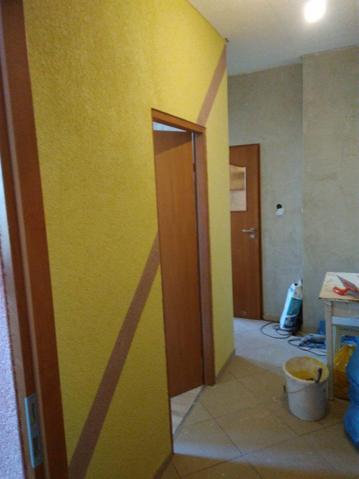 Żółta ściana z dekoracyjnym brązowym pasem, widoczne drzwi wewnętrzne i wiadro z farbą, sugerujące prace malarskie w trakcie realizacji.