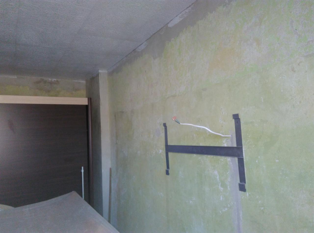 Ściana w trakcie przygotowania do malowania z widocznym uchwytem na telewizor i wystającymi przewodami elektrycznymi, surowy betonowy sufit, widoczne ślady wcześniejszych prac remontowych.
