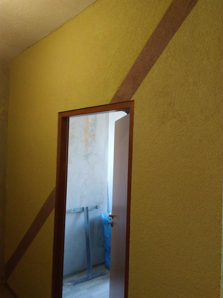 Żółta ściana z dekoracyjnym, ukośnym pasem w odcieniu brązu, widok na otwarte drzwi z widocznym rusztowaniem i workiem wewnątrz pomieszczenia.