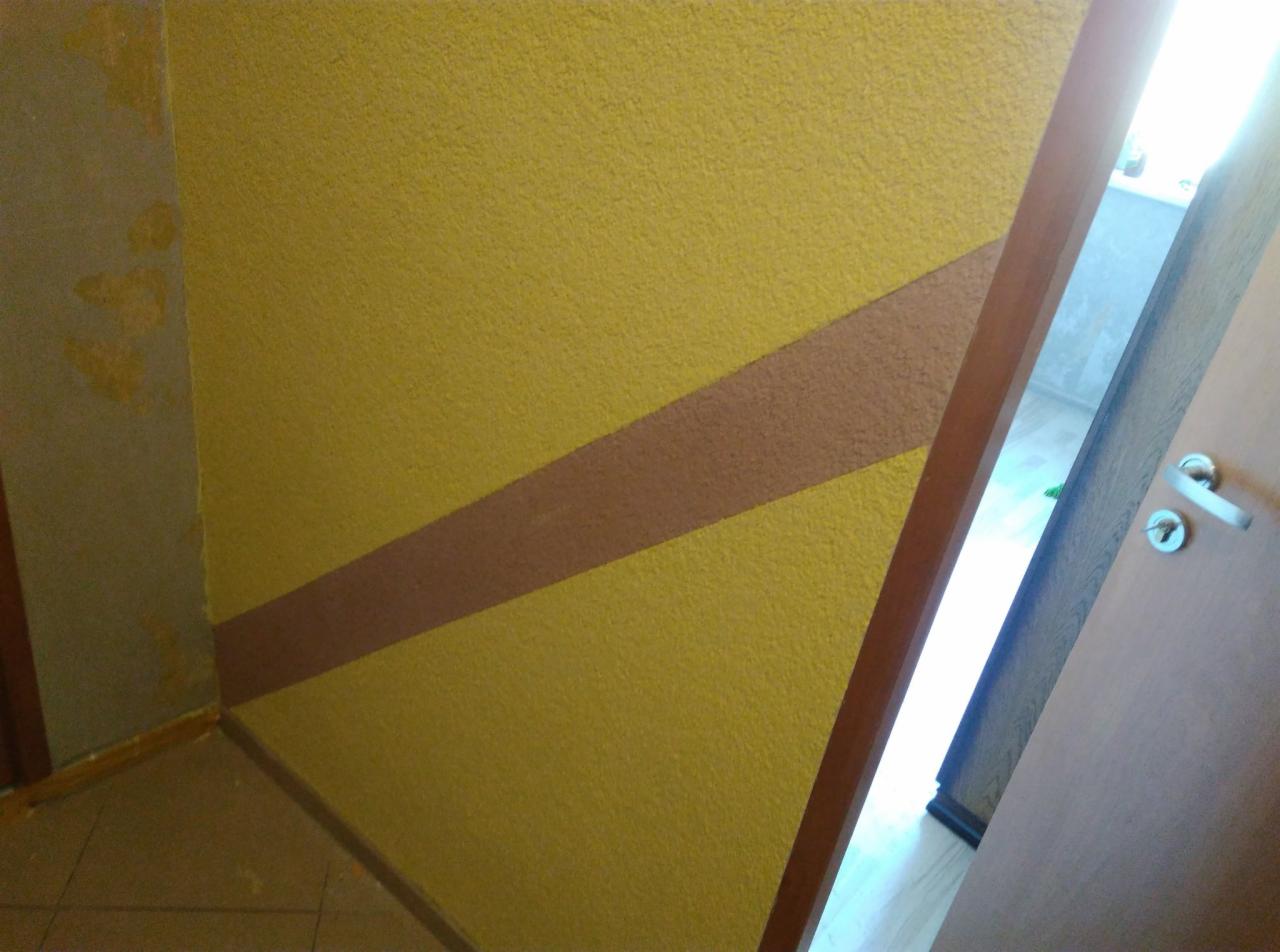 Żółta ściana z teksturą i ukośnym brązowym pasem, widoczny narożnik i fragment drzwi z klamką.